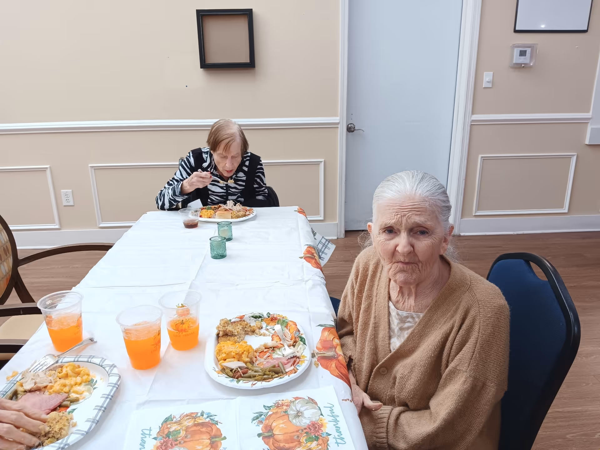 Two elderly women sitting at a long table covered with a white tablecloth and a fall-themed runner, eating a meal with plates of food and cups of orange drink in a dining room with beige walls and wooden floor.
