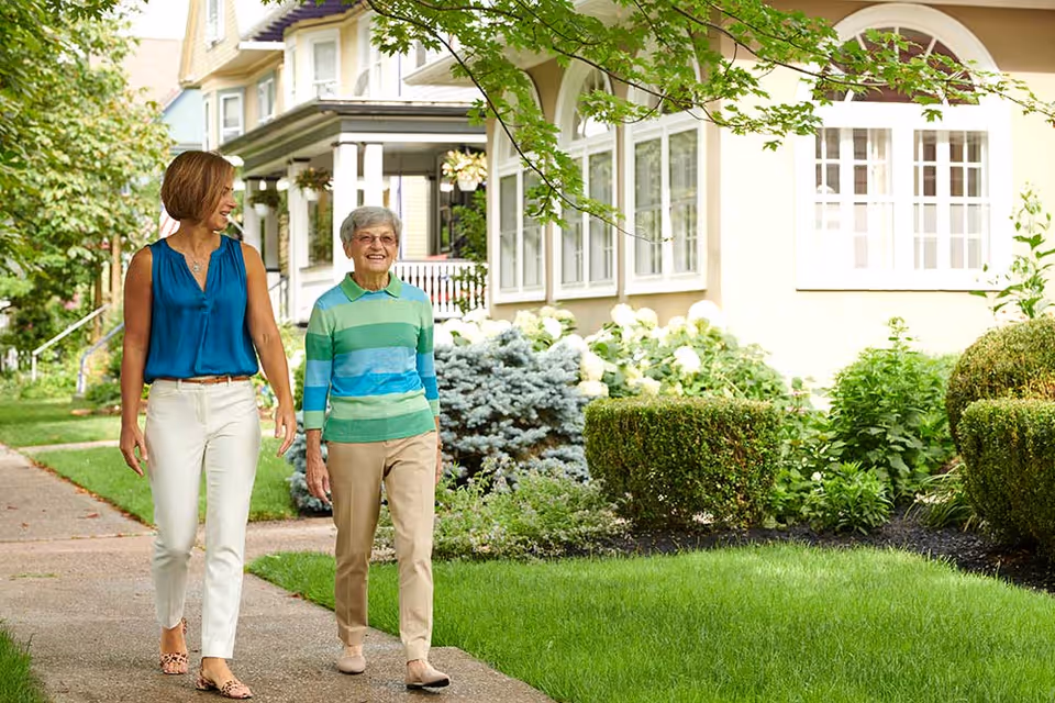 Two women walking and talking on a sidewalk in a residential neighborhood with well-maintained gardens and houses in the background.
