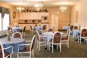 A dining room with several round tables covered with blue tablecloths and surrounded by white chairs with floral cushions. The room has beige walls, two chandeliers, a window with curtains, and a cabinet with decorative items on top.