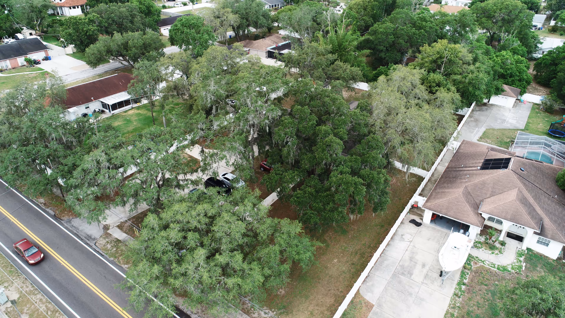 Aerial view of a residential neighborhood with several houses surrounded by many large green trees. A street with a red car driving is visible on the left side. One house has a driveway with a boat parked on it and a fenced backyard with a pool covered by a screened enclosure.