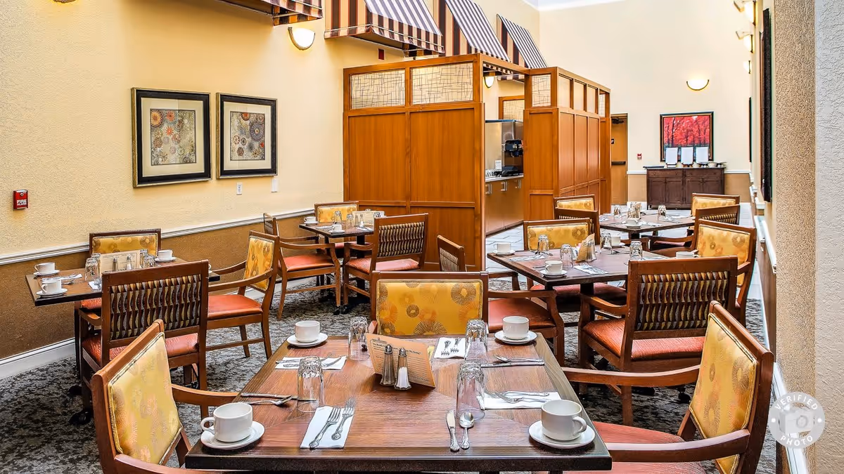 A dining room with several wooden tables and chairs arranged neatly. Each table is set with white cups, glasses, silverware, and napkins. The chairs have yellow and orange patterned cushions. The room has beige walls with framed artwork and striped awnings above a wooden partition that separates the dining area from a kitchen or service area in the background.