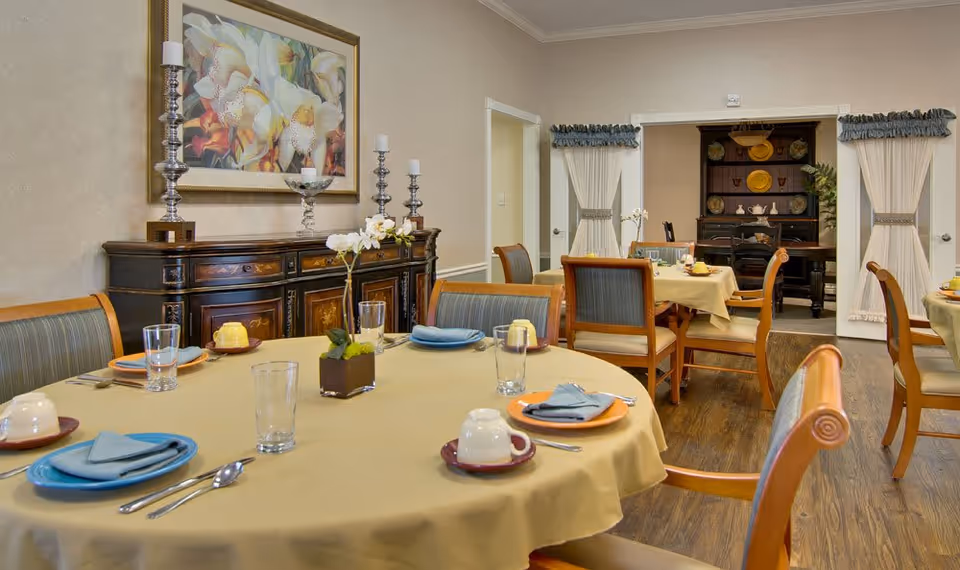 A dining room with round tables covered in beige tablecloths, set with plates, cups, glasses, and silverware. The room has wooden chairs with cushioned seats and backs. A sideboard with decorative candlesticks and a large floral painting hangs on the wall. In the background, there is a dark wooden hutch with plates and teapots displayed, and cream-colored curtains tied back on either side of the doorway.