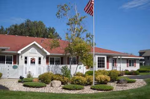Single-story senior living building with red roof, flagpole, and landscaped front yard.