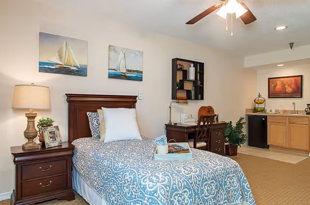 A cozy bedroom featuring a single bed with a blue and white patterned bedspread and a white pillow. Next to the bed is a wooden nightstand with a lamp, a small plant, and a framed photo. Above the bed are two paintings of sailboats on the water. In the background, there is a wooden desk with a chair, a wall-mounted shelf, a potted plant, and a small kitchenette area with a sink, mini fridge, and cabinets. The room has beige walls and carpeted flooring, with a ceiling fan overhead.