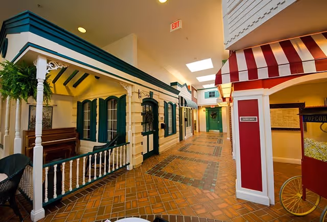Indoor hallway designed to look like a small town street with storefront facades, including a porch with a piano, green doors and windows, a red and white striped awning, and a popcorn cart on the right side. The floor is tiled with a brick pattern and the ceiling has skylights.