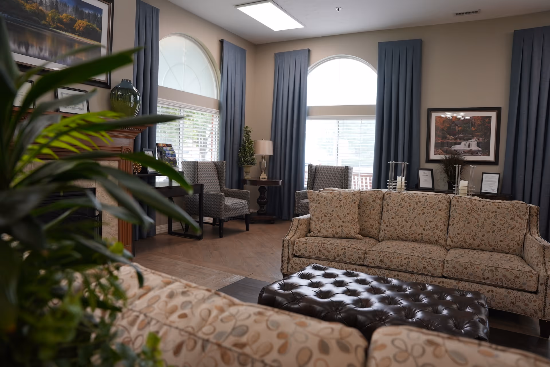 A cozy living room with patterned beige sofas, a dark tufted leather ottoman, two armchairs near large arched windows with blue curtains, a side table with a lamp, framed artwork on the walls, and a green plant in the foreground.