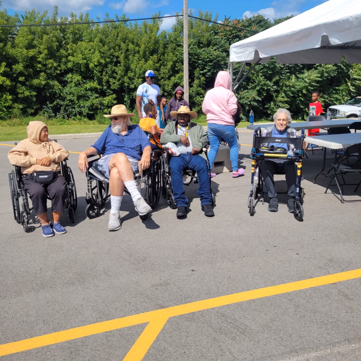 A group of elderly individuals, some in wheelchairs and one using a walker, sitting and socializing outdoors on a sunny day. Behind them, a few people stand near a white canopy tent and folding tables. Green trees and bushes form the background.