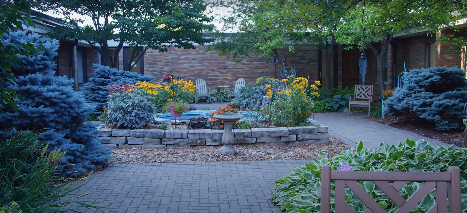A peaceful outdoor garden area with a paved walkway leading to a raised flower bed filled with colorful flowers and greenery. There are several wooden chairs placed around the garden, surrounded by trees and shrubs, with a brick building wall in the background.