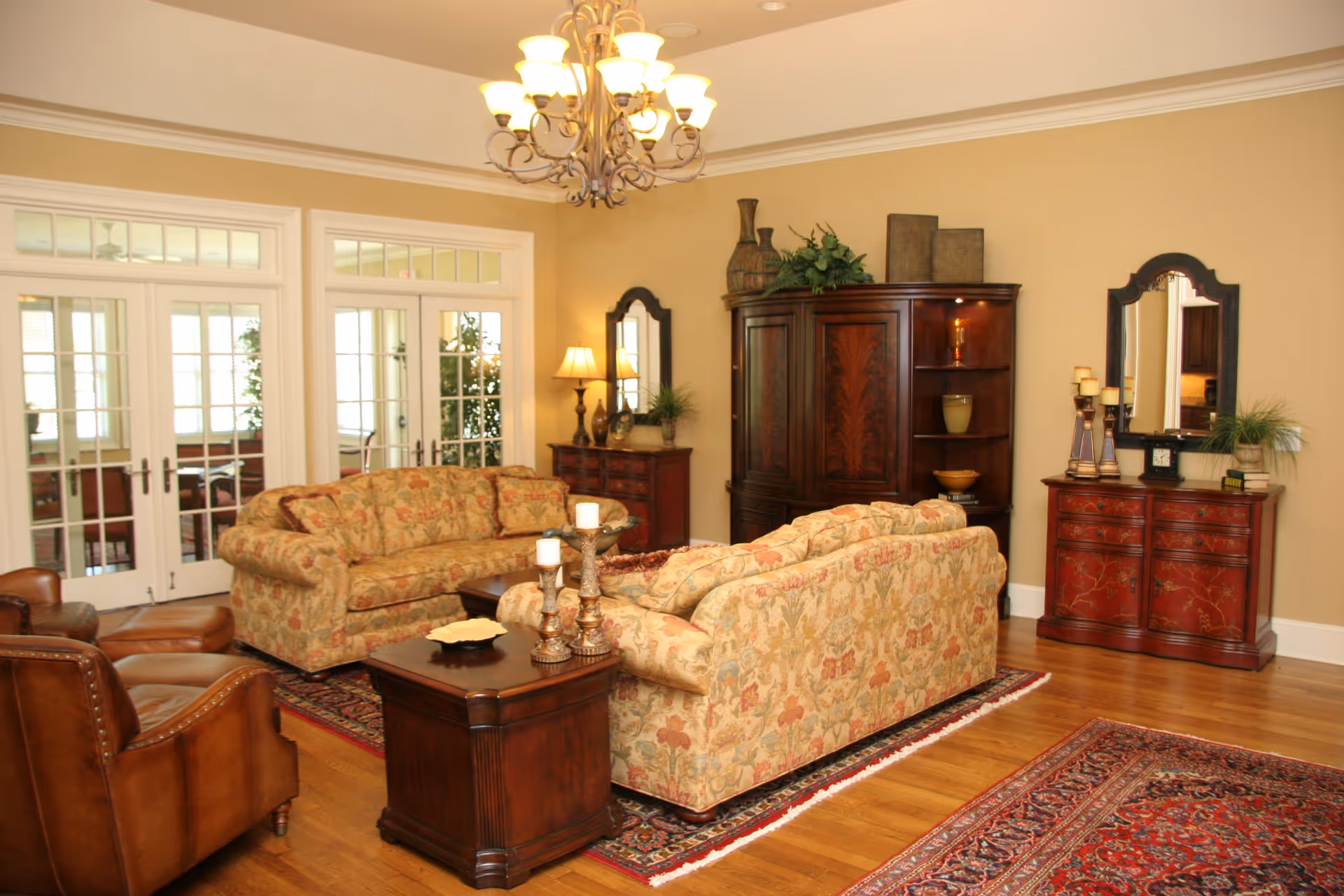 A cozy living room with two floral patterned sofas, a brown leather armchair, a wooden coffee table with candles, and a wooden cabinet with decorative items. The room has hardwood floors, two red patterned rugs, beige walls, a chandelier, and French doors with glass panes leading to another room.