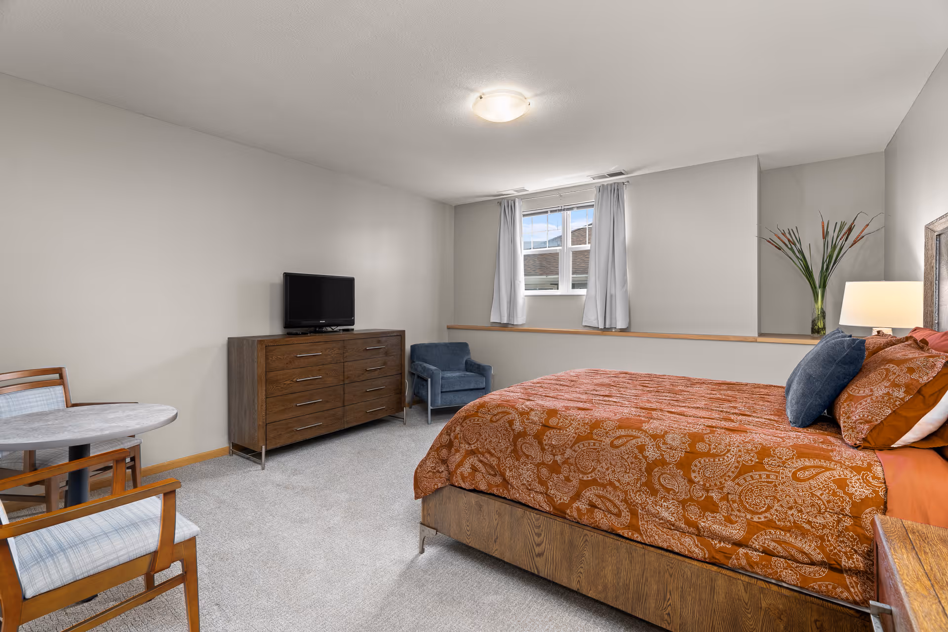 A bedroom with a bed covered in an orange patterned bedspread, a wooden dresser with a TV on top, a blue armchair, a small round table with two chairs, a window with white curtains, and a vase with tall flowers on a shelf.