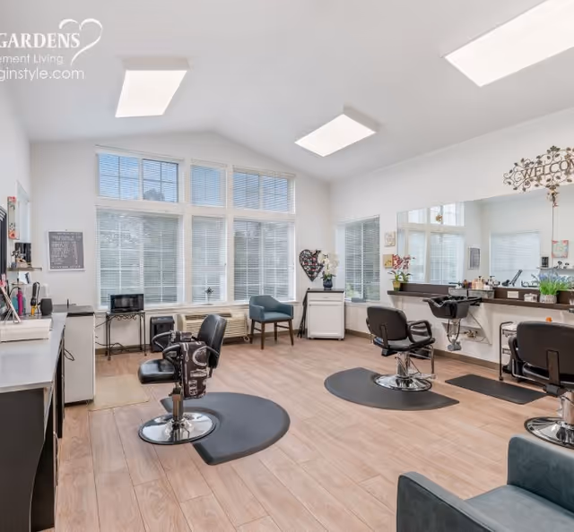 A bright and clean salon area with three black salon chairs on black mats, a large mirror on the wall, and a washing station. The room has large windows with blinds, light wood flooring, and white walls. There are some plants and decorative items on the counters and a blue armchair near the windows.