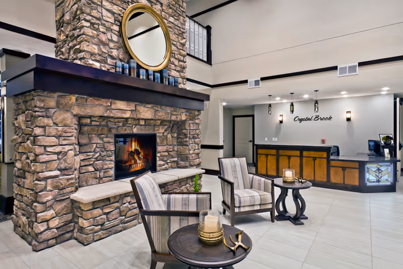 Interior view of a senior living facility lobby featuring a large stone fireplace with a round mirror above it, two striped armchairs, and two small round tables with candles. In the background, there is a reception desk with the sign 'Crystal Brook' on the wall behind it.