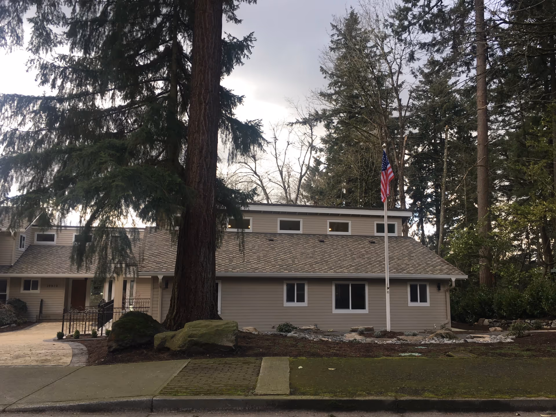 Front exterior of a one-story house-like building with an American flag and large trees in front.