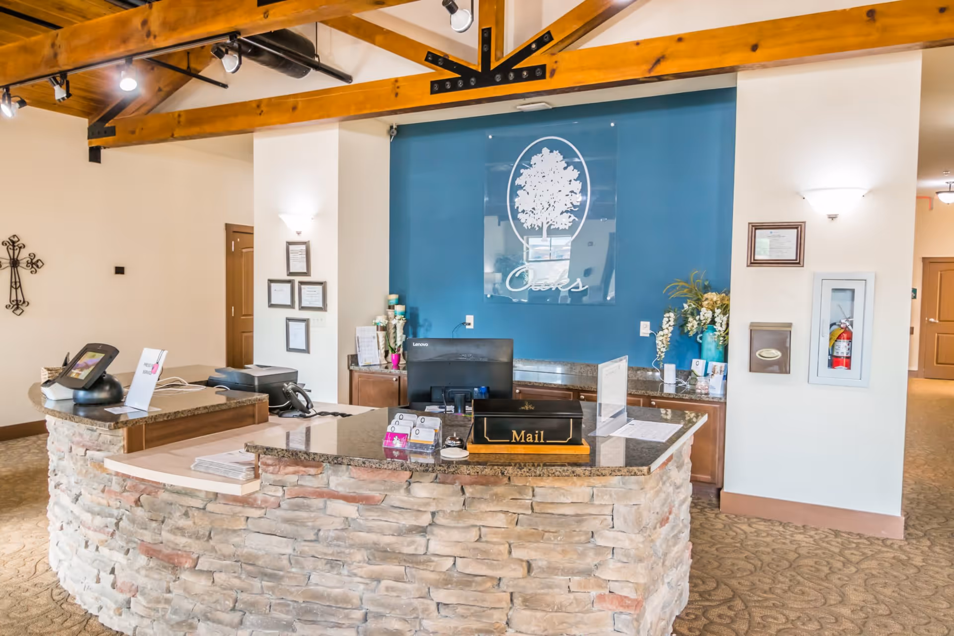 Reception desk area inside Oaks at Pooler facility with a stone front counter, computer, mail box, and decorative items. Behind the desk is a blue wall with a large logo featuring a tree and the word 'Oaks'. The ceiling has exposed wooden beams and track lighting.