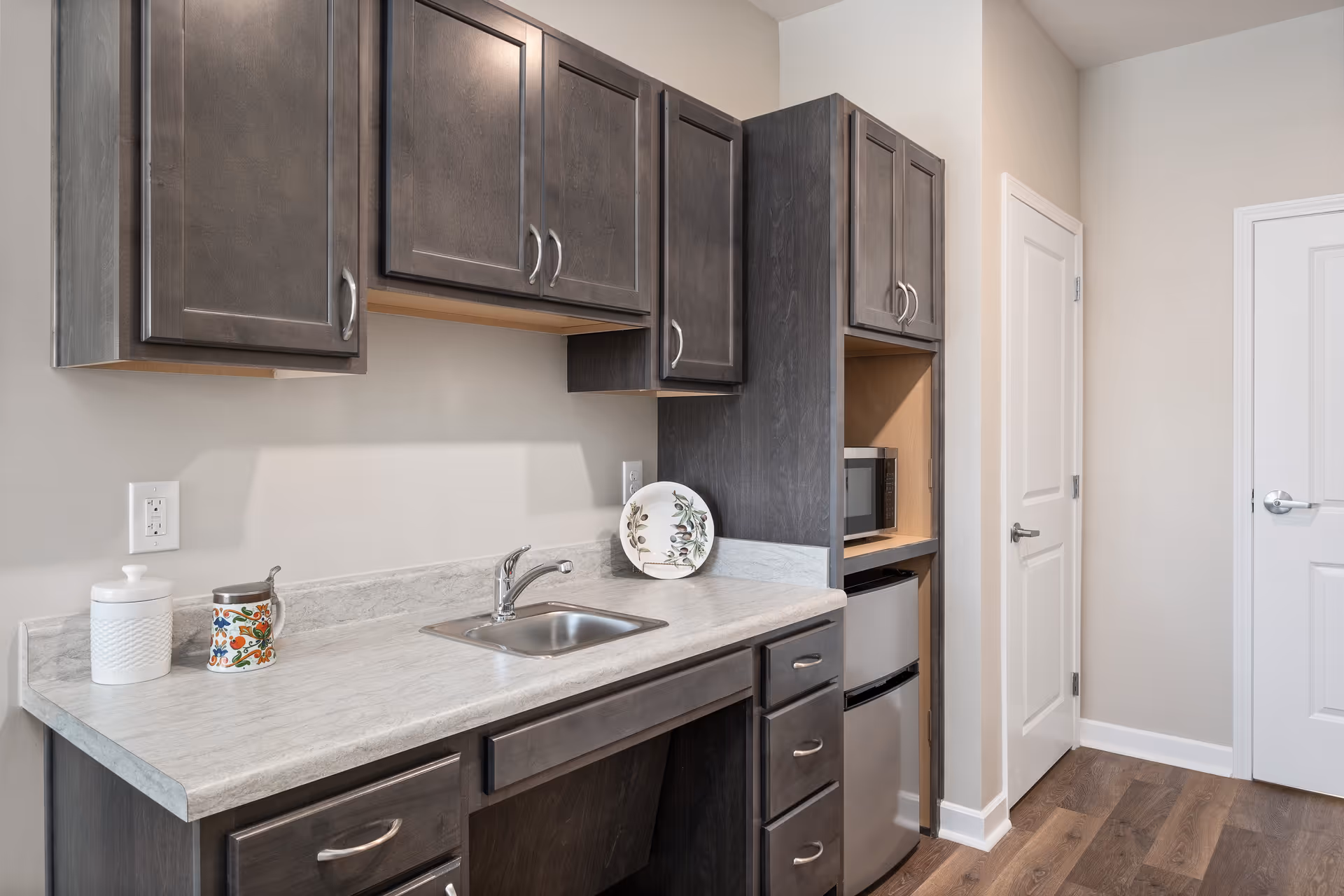 A small kitchen area with dark wood cabinets, a light-colored countertop with a built-in sink, a mini refrigerator, and a microwave. There are decorative items on the counter including a white jar, a colorful mug, and a decorative plate. The floor is wood, and there are two white doors in the background.