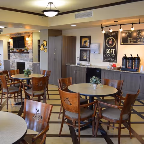 A cozy dining area with round tables and wooden chairs featuring coffee cup designs on the backs. The room includes a kitchenette with a countertop, sink, coffee dispensers, and a chalkboard sign advertising soft serve and coffee options. There is a fireplace with a mounted TV above it in the background, and the floor has a checkered pattern.