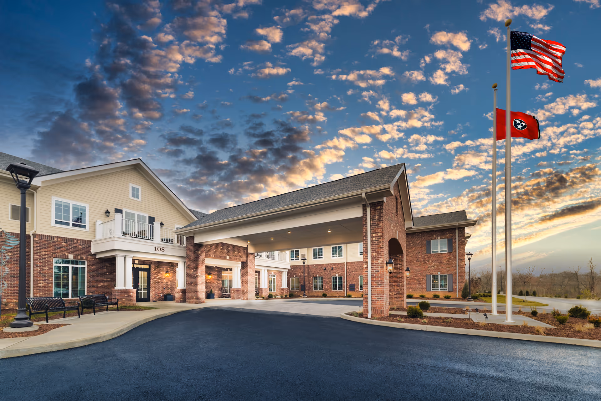 Front entrance of a brick senior living building with a covered porte-cochere, flagpoles, benches, and a dramatic cloudy sky.