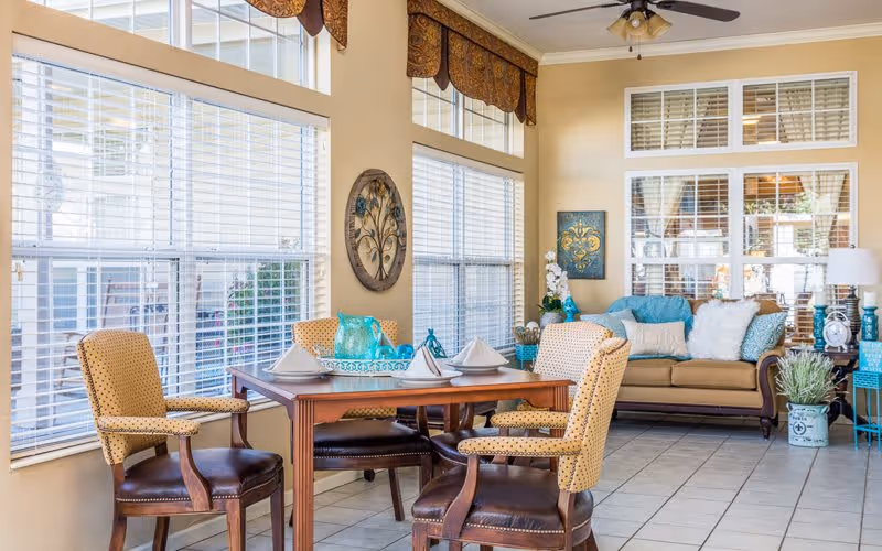 A bright and cozy interior space featuring a wooden dining table set with four upholstered chairs, each with a yellow patterned fabric. The table is set with white napkins and turquoise decorative items. Large windows with white blinds allow natural light to fill the room. In the background, there is a beige sofa adorned with blue and white cushions, a side table with a lamp, and decorative wall art. The floor is tiled, and the ceiling has a fan with lights.