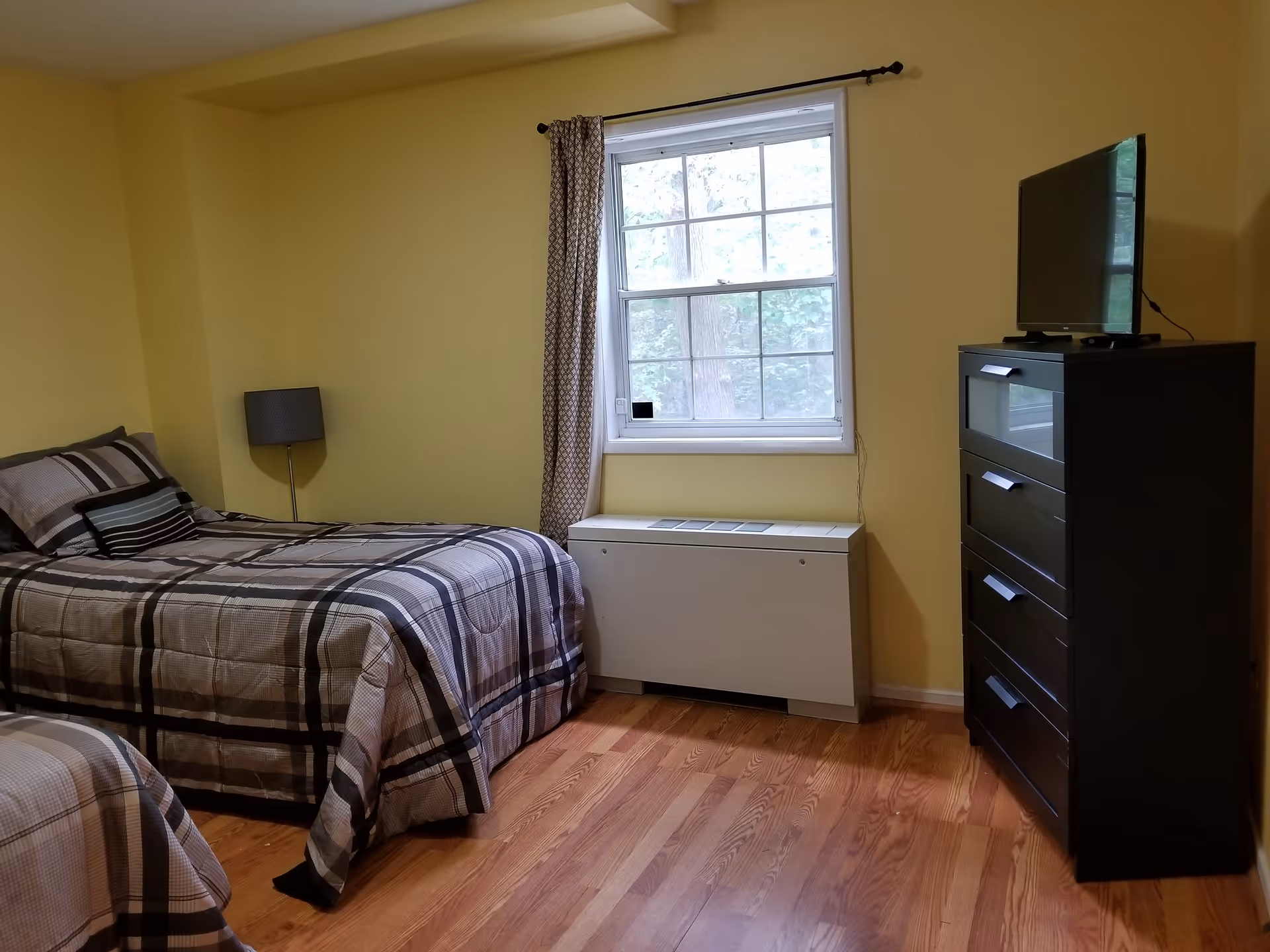 A bedroom with yellow walls featuring two single beds with plaid bedding, a floor lamp between the beds, a window with patterned curtains, a white radiator below the window, and a black dresser with a flat-screen TV on top.