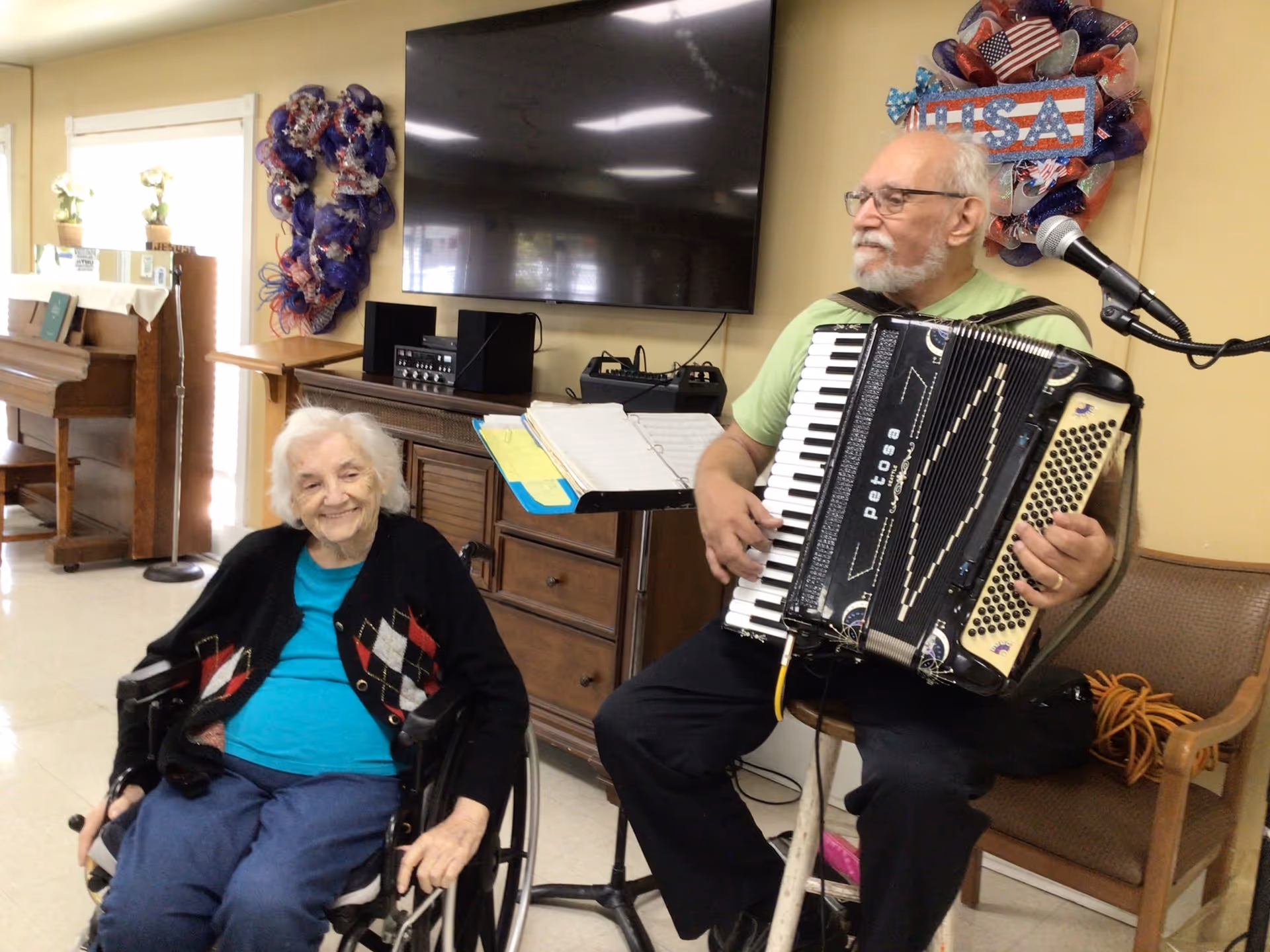 An elderly woman in a wheelchair smiling while a man plays an accordion next to her in a room decorated with patriotic wreaths and a large TV mounted on the wall. There is a piano and a music stand with sheet music in the background.