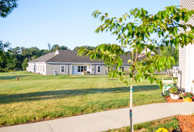 View of a single-story building with beige siding and a dark roof, surrounded by a well-maintained grassy lawn and a few small trees. A concrete sidewalk runs along the foreground, and there is a garden hose and some potted plants near the building on the right side.