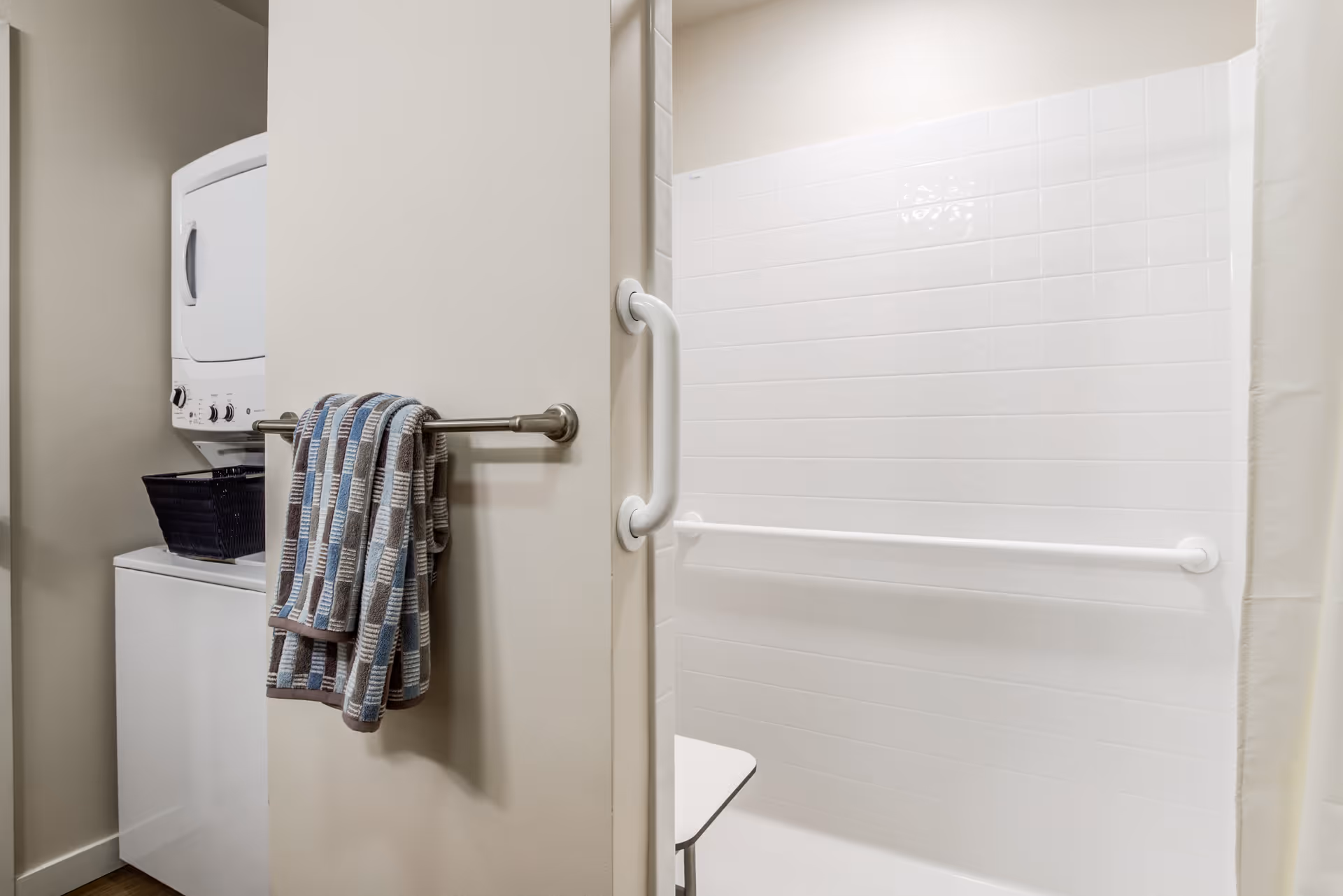 A bathroom area with a white tiled shower featuring grab bars and a foldable shower seat. Adjacent to the shower is a laundry area with a stacked washer and dryer, and a towel hanging on a metal towel rack.