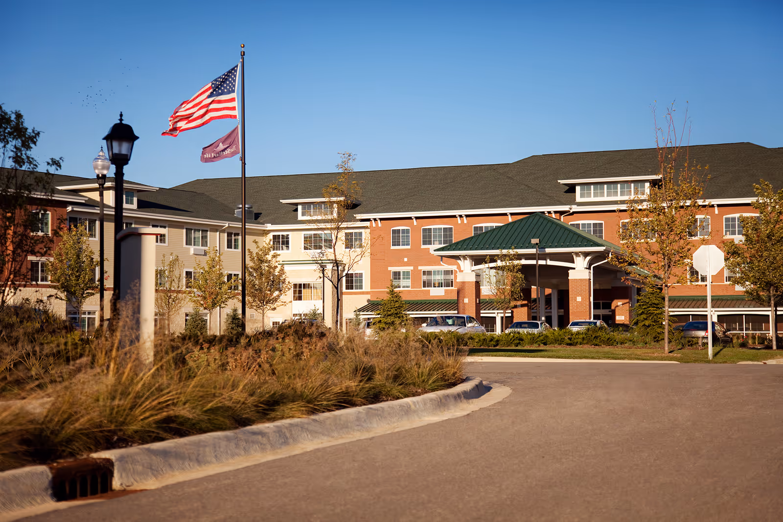Exterior view of a senior living facility building with a green roof entrance canopy, multiple windows, and a driveway. There are two flagpoles with the American flag and another flag, trees, and street lamps along the driveway under a clear blue sky.