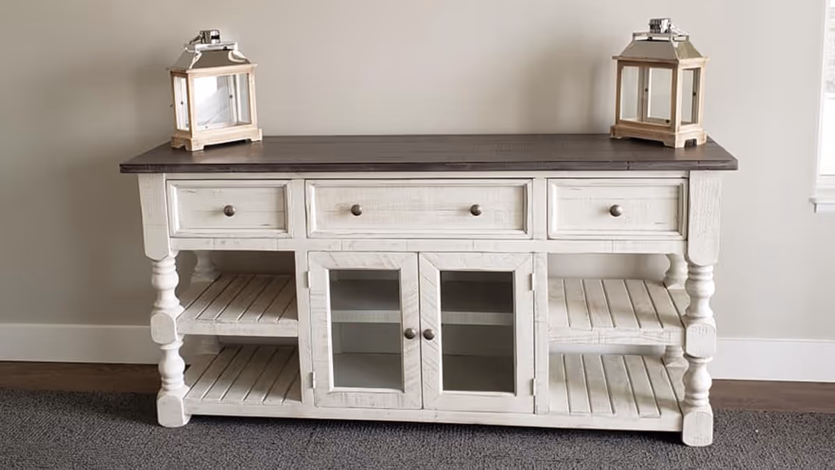 White distressed wooden console table with a dark top and two lanterns on it against a neutral wall.