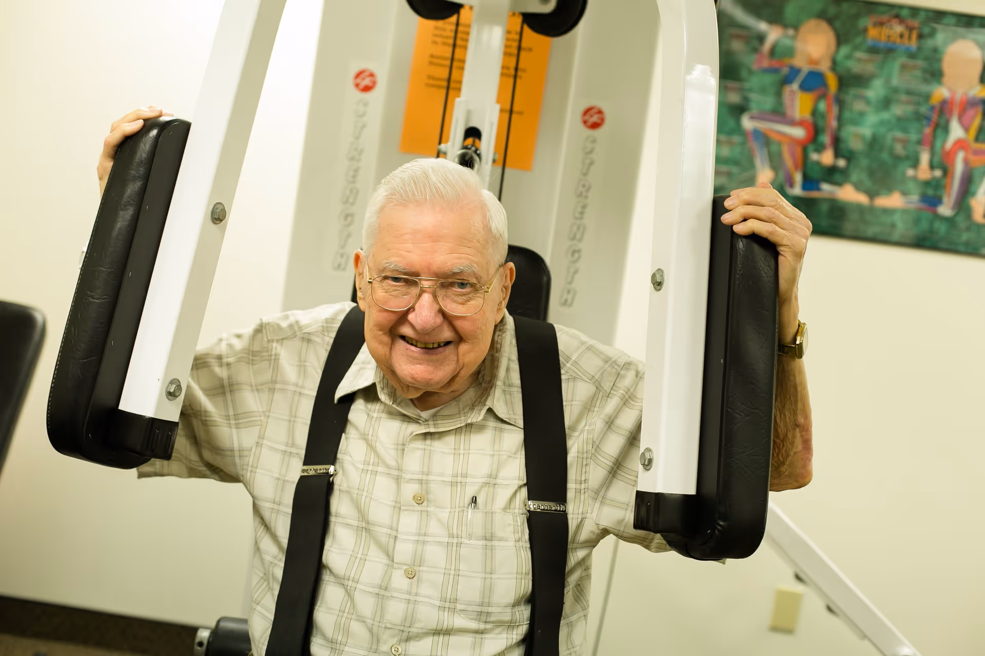An elderly man with white hair and glasses is smiling while using a chest press exercise machine in a fitness room. He is wearing a plaid shirt with black suspenders. There is a colorful anatomical poster on the wall behind him.