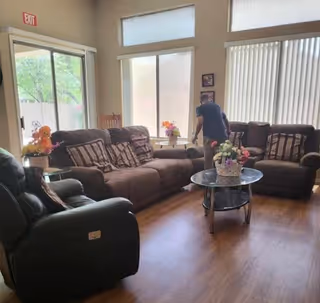 A bright common living room with sofas and recliners around a glass coffee table topped with flower arrangements, and a person standing by the windows.