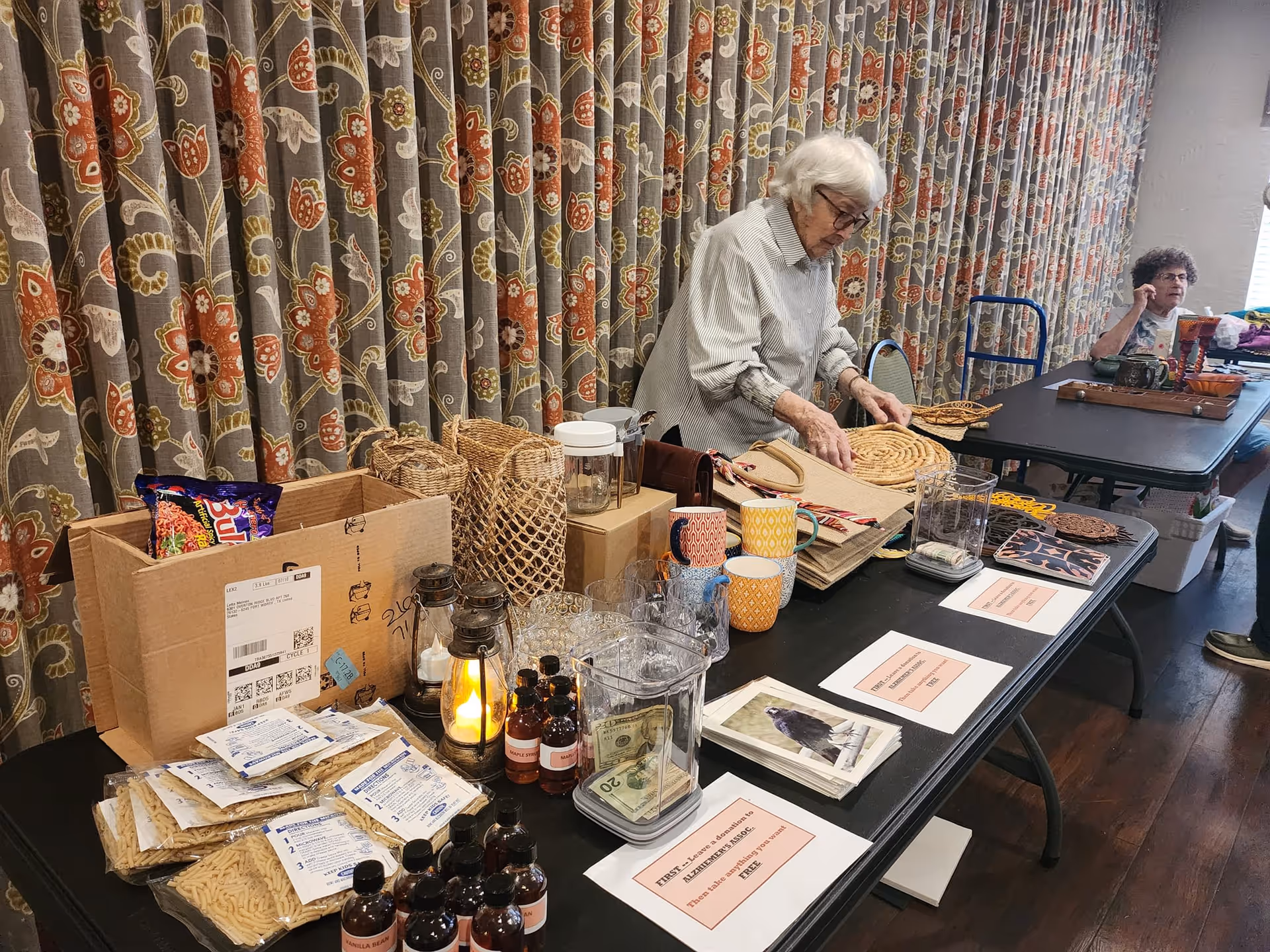 An elderly woman arranging woven placemats and other items on a long table covered with various objects including mugs, glassware, packets of food, and informational signs. Another elderly woman is seated at the far end of the table near a window. The background features floral patterned curtains.