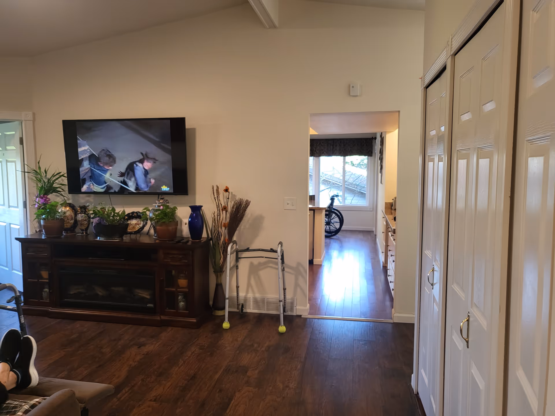 A living room area with a mounted flat-screen TV showing a video, a wooden cabinet with plants and decorative items, a walker with tennis balls on the legs, and a view into a kitchen area with a wheelchair near a window.