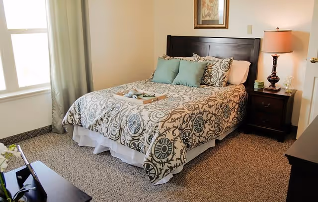 A neatly made bed with patterned bedding and three decorative pillows in a bedroom. There is a wooden nightstand with a lamp and small decorative items next to the bed. A window with light curtains allows natural light into the room. The floor is carpeted and there is a framed picture hanging on the wall above the bed.