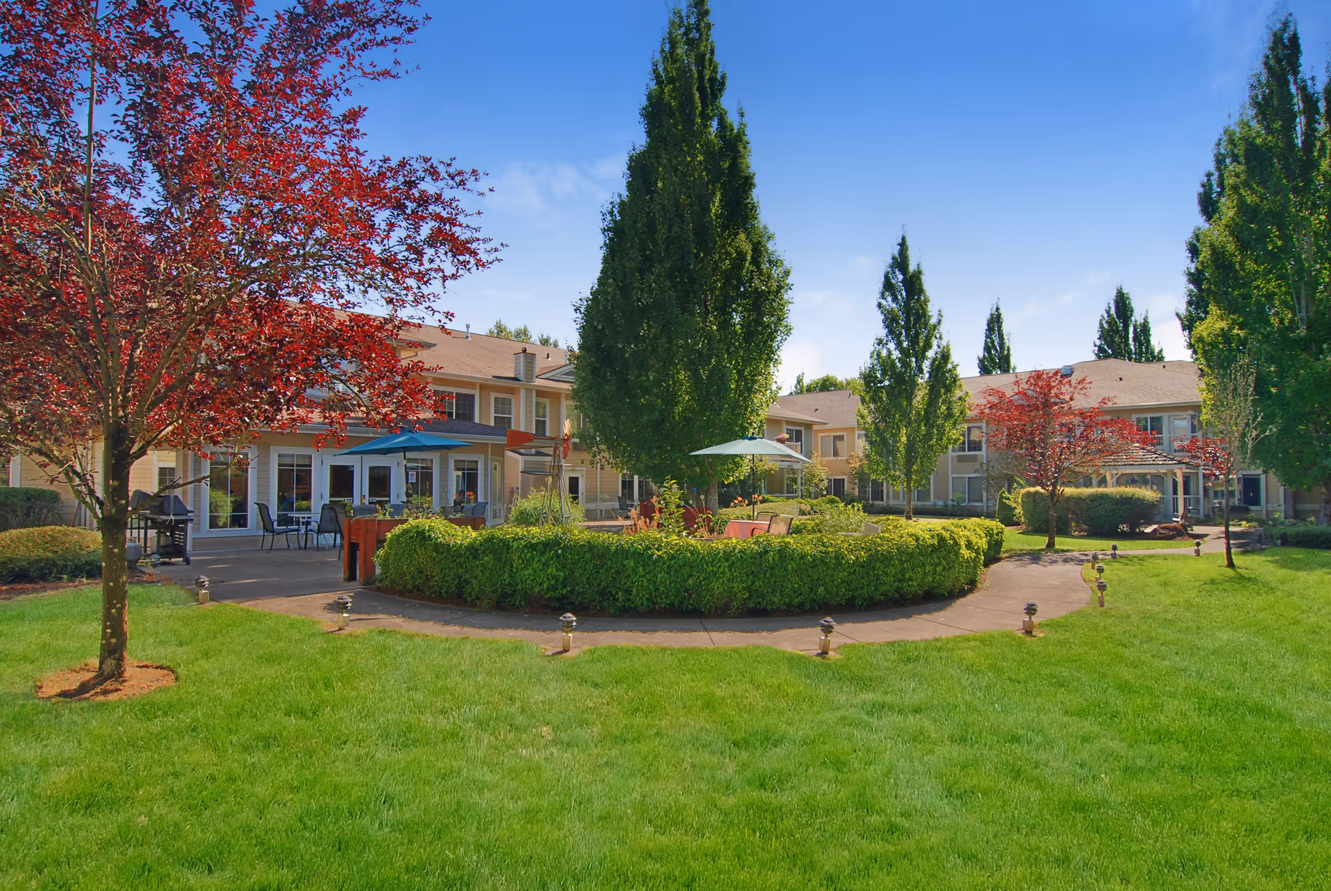 Sunlit landscaped courtyard with seating areas, umbrellas, trees and a two-story residential building in the background.