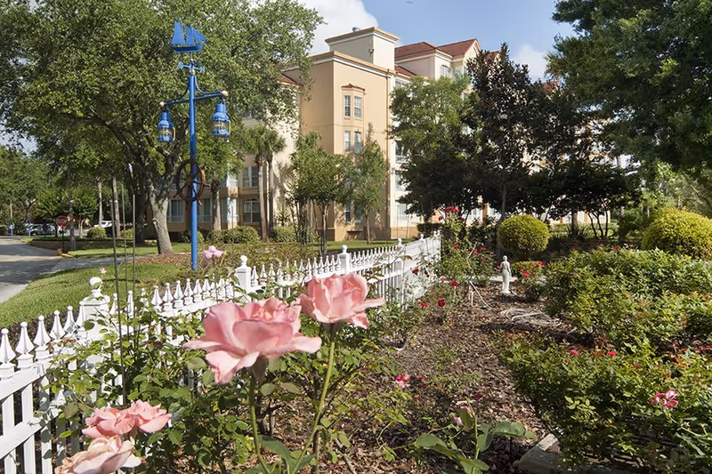 A landscaped garden area with pink roses and various green shrubs bordered by a white picket fence. In the background, there is a multi-story beige building partially obscured by trees. A blue lamp post with nautical-themed decorations is visible near the fence.