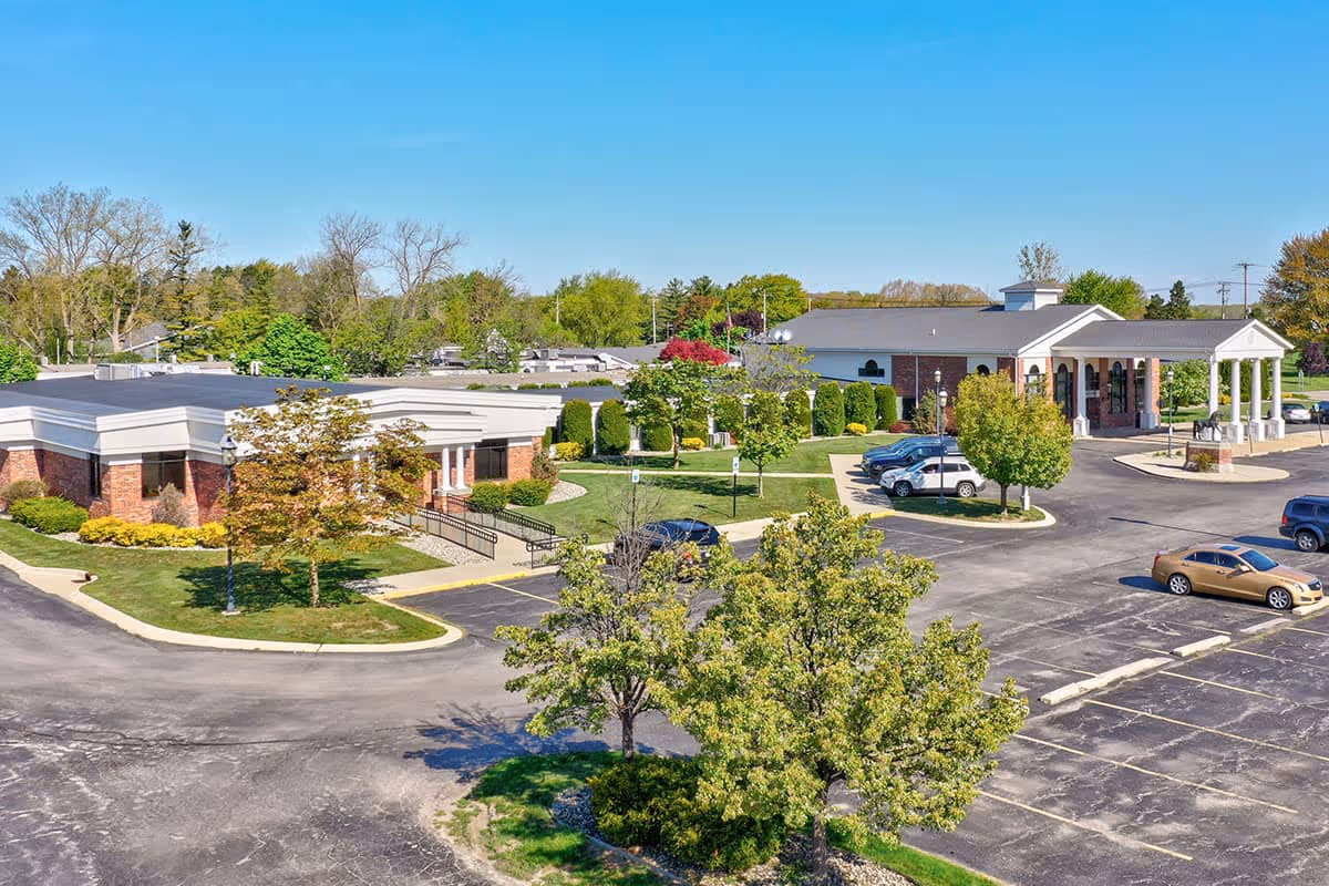 Aerial view of a nursing home complex with landscaped lawns, trees, parking lot, and a main entrance with a covered portico.
