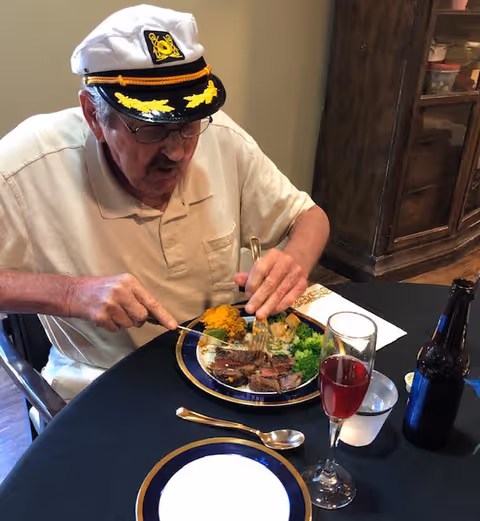 An elderly man wearing a white captain's hat and glasses is sitting at a table cutting food on a plate. The plate contains steak, broccoli, mashed sweet potatoes, and other vegetables. On the table, there is a glass of red beverage, a bottle, a spoon, and a napkin. The setting appears to be indoors with a wooden cabinet in the background.