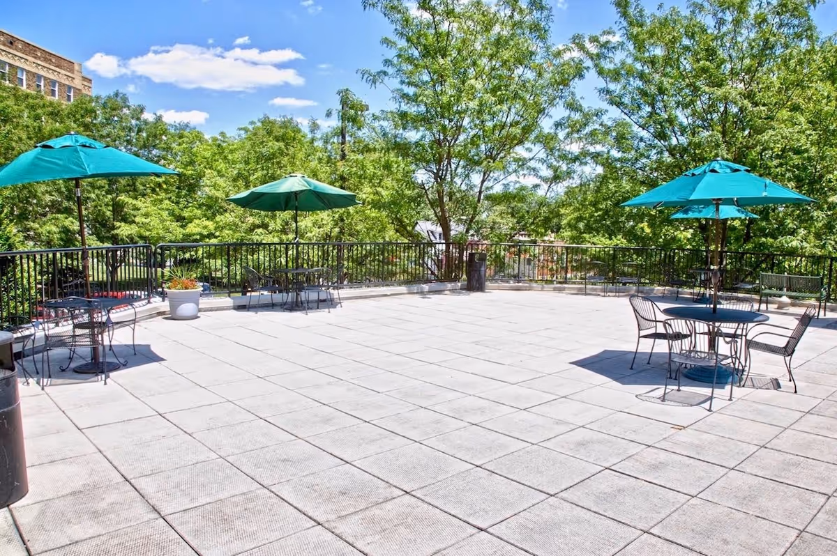 Outdoor patio area with tiled flooring, several metal tables and chairs, each shaded by green umbrellas, surrounded by trees and greenery under a blue sky with some clouds.
