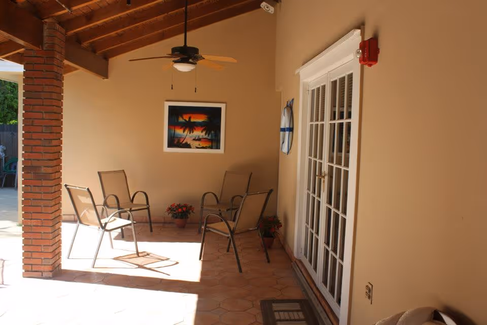 Covered outdoor patio area with four metal-framed chairs arranged around a small space, a ceiling fan with wooden blades hanging from a wooden beam ceiling, a framed tropical sunset painting on the beige wall, two potted plants on the floor, and a set of white French doors on the right side.