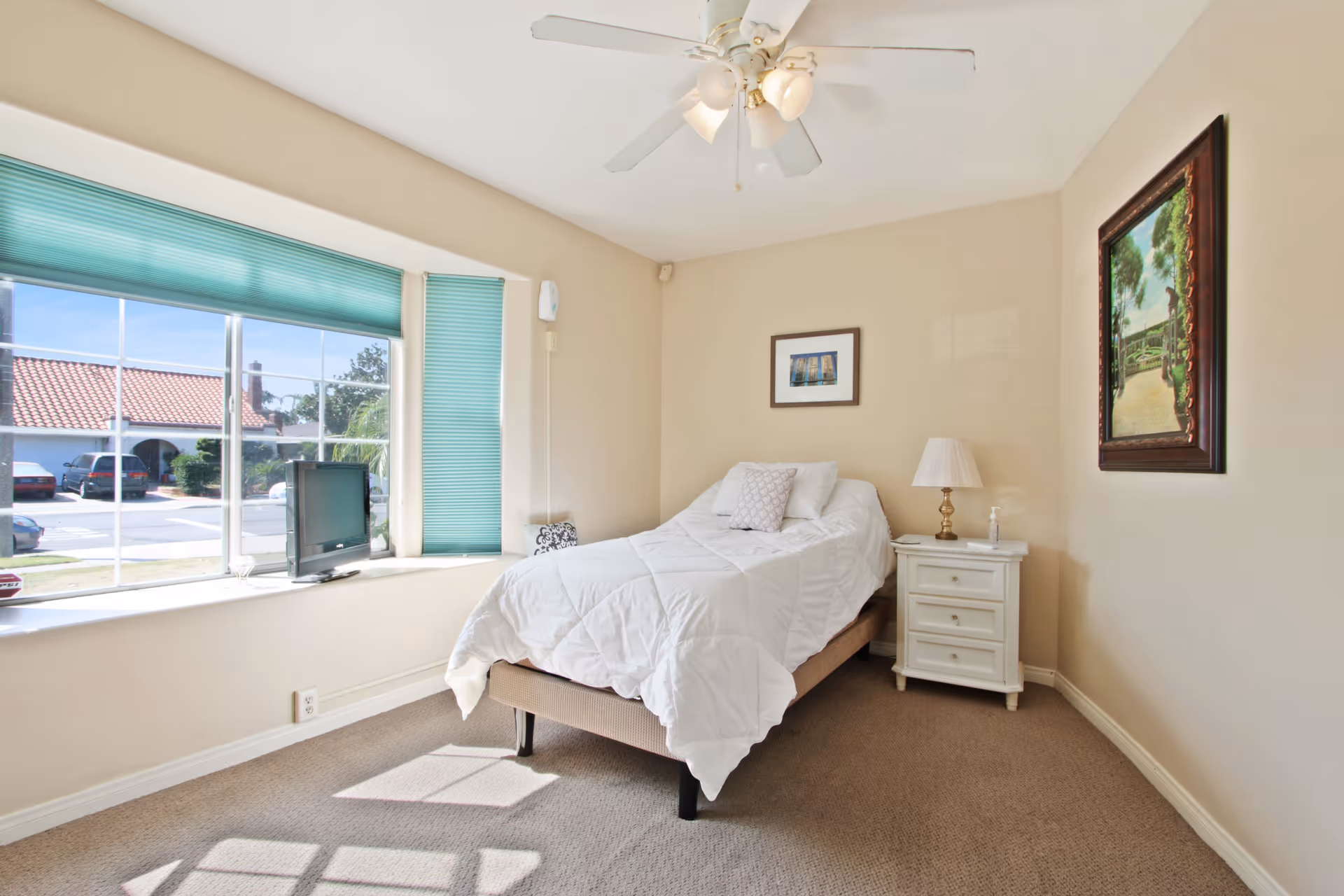 A bright bedroom with a single bed covered in white bedding, a white nightstand with a lamp and a bottle of hand sanitizer, a ceiling fan with lights, a large window with turquoise blinds letting in natural light, and two framed pictures on the beige walls.