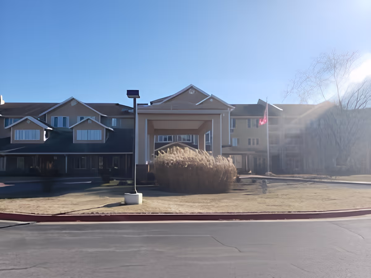 Front entrance of a multi-story senior living building with a covered porte-cochère, flagpole, and landscaped lawn.