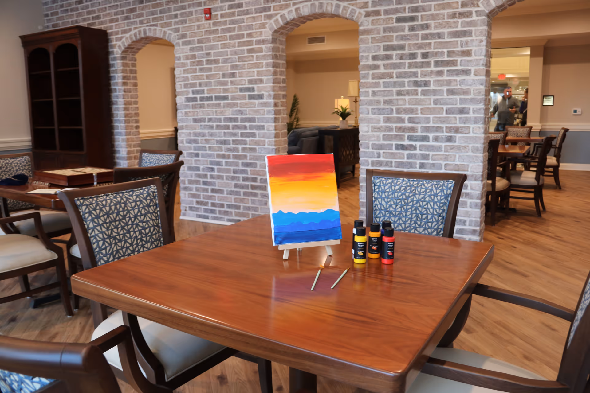 A wooden table with four chairs around it in a room with brick archways. On the table is a small canvas on an easel featuring a colorful painting of a sunset over mountains, along with three bottles of paint and two paintbrushes. The room has wooden flooring and additional tables and chairs in the background.