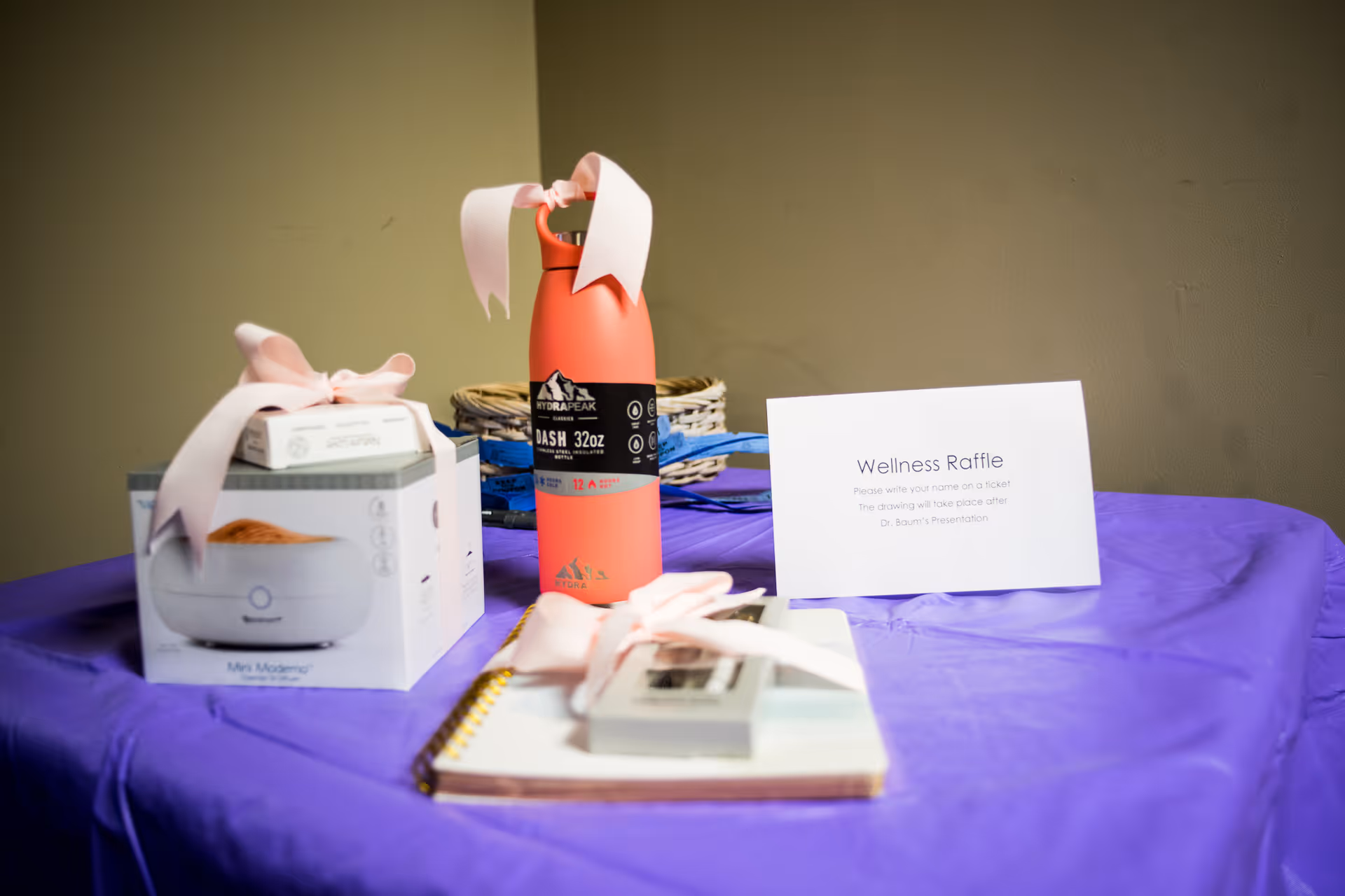 A table covered with a purple cloth displaying raffle prizes including a pink water bottle, a white boxed item with a pink ribbon, and a notebook with a pink ribbon. A sign on the table reads 'Wellness Raffle Please write your name on a ticket The drawing will take place after Dr. Baum's Presentation.'