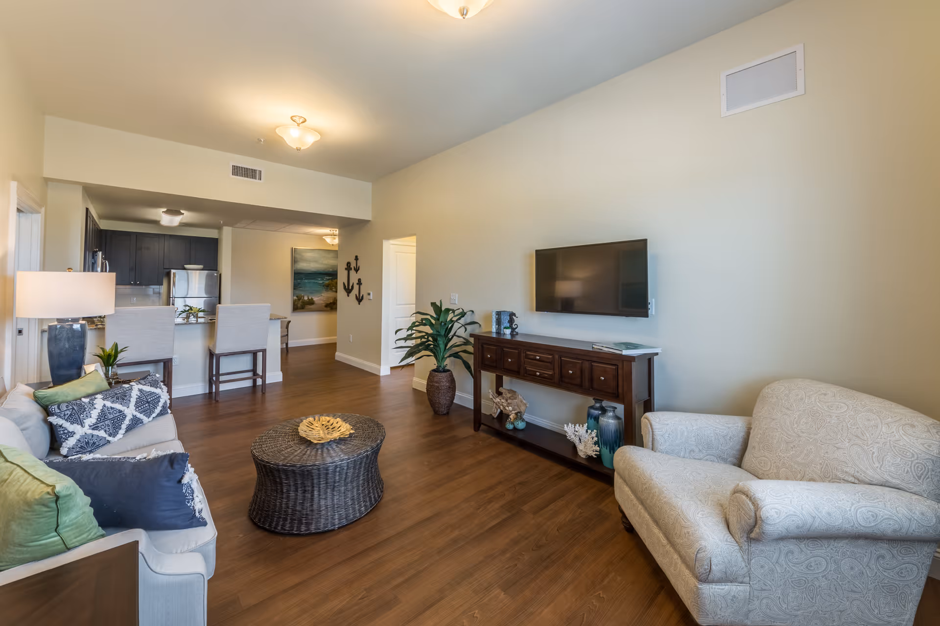 A bright and spacious living room area with wooden flooring, a white patterned armchair, a white sofa with green and blue cushions, a round wicker coffee table, a wooden console table with decorative items, and a wall-mounted flat-screen TV. The room opens into a kitchen area with a breakfast bar and stools, and there is a hallway with wall art and anchor decorations.