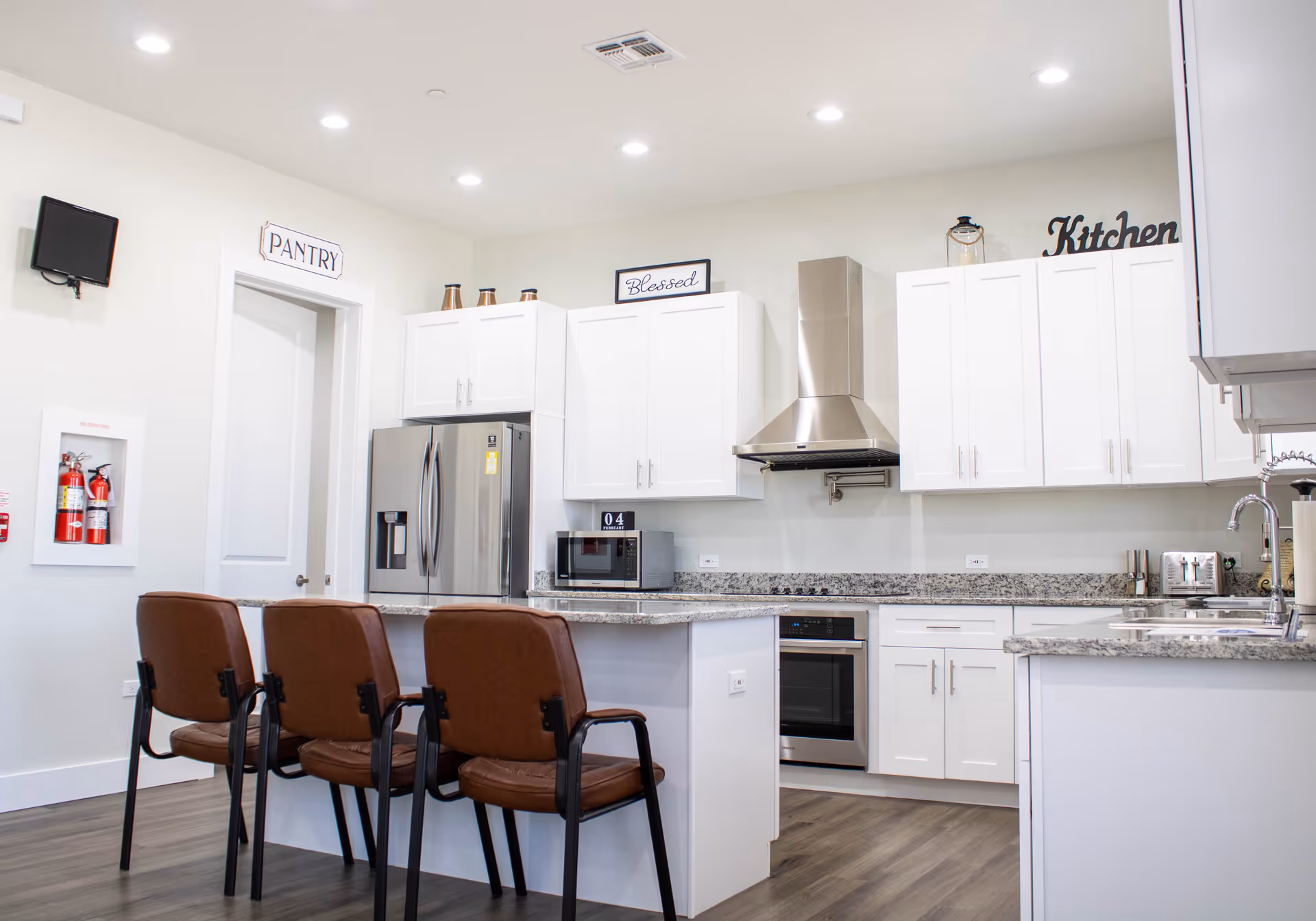 Bright modern communal kitchen with white cabinets, granite countertops, stainless steel appliances and three brown barstools at an island.