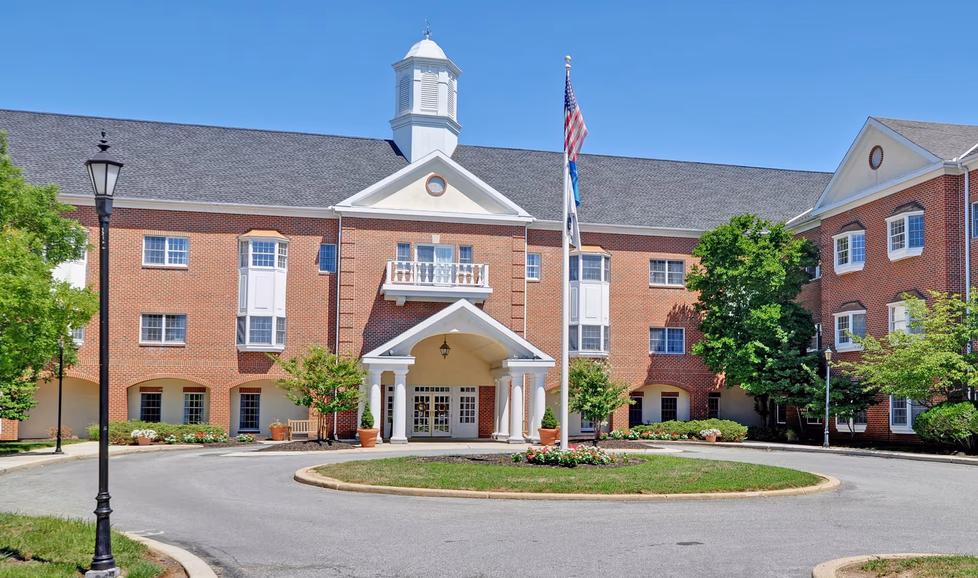 Front exterior view of a large brick building with white trim, a covered entrance supported by columns, a circular driveway with a flagpole displaying the American flag and other flags, and landscaped greenery including trees and flower beds under a clear blue sky.