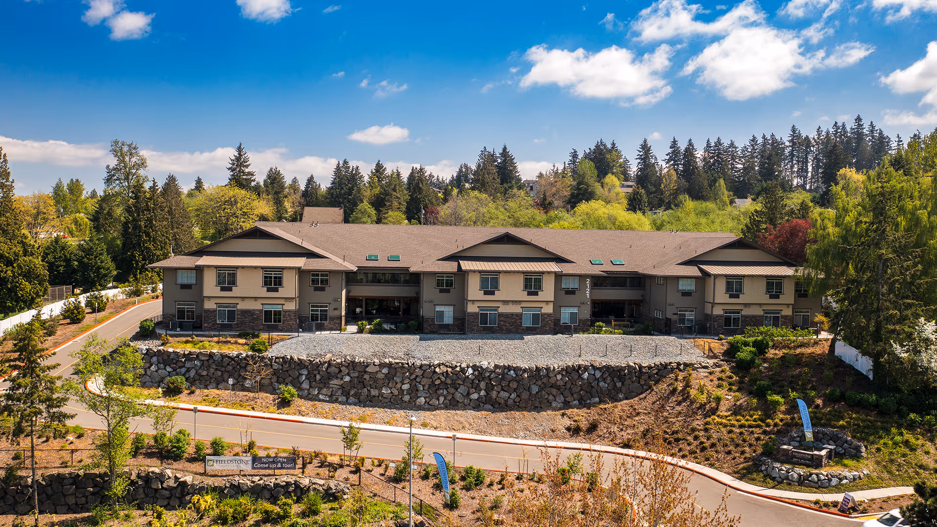 Exterior view of Fieldstone Memory Care Puyallup, a two-story building surrounded by trees and landscaping under a blue sky with some clouds. The building has a stone retaining wall in front and a driveway leading up to it.