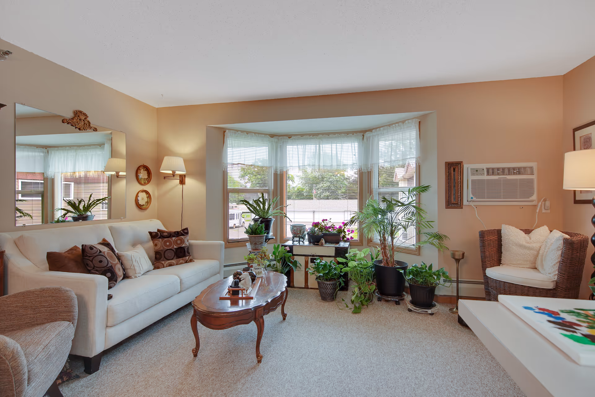 Cozy living room with a sofa, coffee table, armchairs, a bay window filled with potted plants, and a wall air conditioner.