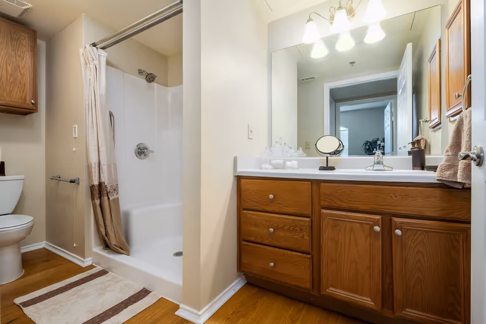 A clean bathroom featuring a white toilet, a shower with a beige and brown curtain, a wooden vanity with a white countertop, a large mirror, and overhead lights. The floor is wooden with a beige and brown striped rug in front of the shower.