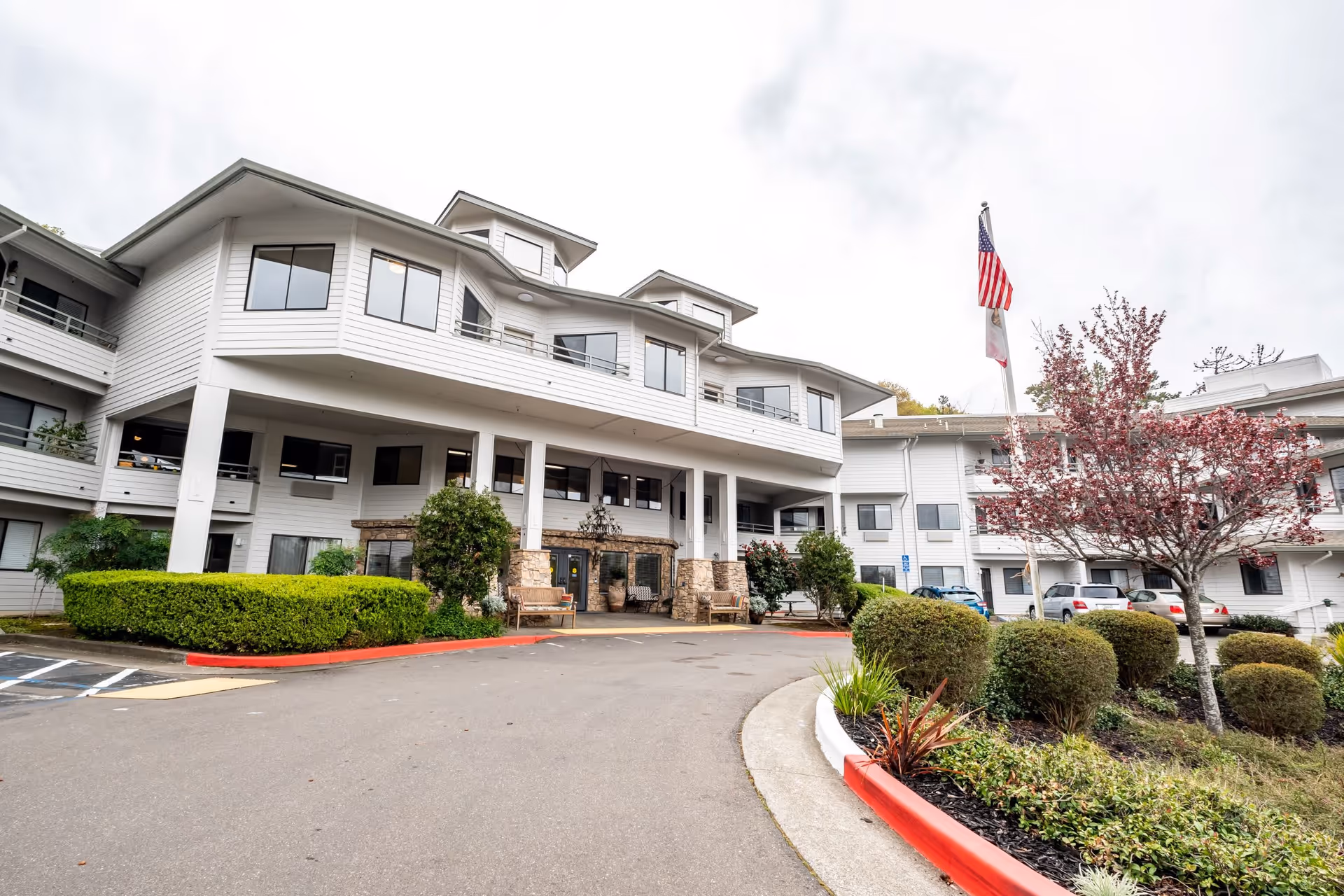 Exterior view of a multi-story white retirement community building with large windows, balconies, and a covered entrance. There are landscaped bushes, a tree with red leaves, and an American flag on a flagpole near the entrance. Several parked cars are visible in the background.